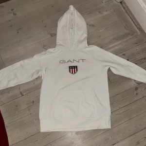 Vit hoodie från GANT med broderad logga - Säljer en vit hoodie från GANT med broderad logga och sköld på bröstet. Tröjan har huva, lång ärm och en stor magficka framtill. Klassisk och stilren design med sportig känsla. Inte urtvättat men de sista bild en liten skrapa som nog går bort i större tvätt men inte haft orken att göra det själv därav priset. 