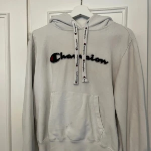 Vit hoodie från Champion - Vit hoodie från Champion med broderad logga på bröstet och tryck på snörena. Klassisk känguruficka framtill och ribbade muddar. Perfekt för en avslappnad stil. Har dock en liten fläck på fickan som ej går bort men syns inte så mycket