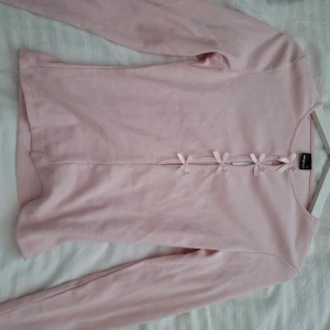 Ljuslila ribbad topp med rosetter från Gina Tricot - Supersöt ljusrosa långärmad topp från Gina Tricot med ribbad struktur och dekorativa rosetter framtill. Perfekt för dig som gillar gulliga detaljer och en feminin stil. Modellen är figurnära och mjuk mot huden.