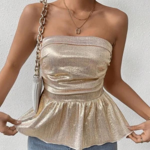 Guldig glittrig tubtopp med volang - Säljer en snygg guldig tubtopp med glittrigt tyg och volang nertill. Toppen är axelbandslös och har en figurnära passform upptill som blir vidare nedtill. Perfekt för att sticka ut på festen!