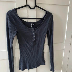 Grå/Blå ribbad långärmad topp från Pieces - Säljer en grå/blå långärmad topp från Pieces med ribbad struktur och knappar framtill. Toppen har en djup rundringning och är figurnära i modellen. Perfekt att styla med jeans eller kjol.