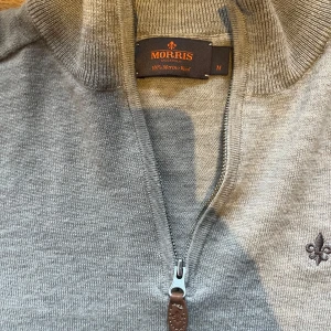 Ljusgrå merinoullströja med dragkedja från Morris - Säljer en ljusgrå tröja från Morris i 100% merinoull. Tröjan har halv dragkedja framtill, ribbade muddar och en liten broderad logga på bröstet. Den är storlek M men passar S bra också.