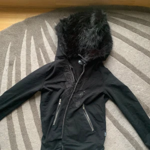 Svart hoodiejacka med fuskpäls och broderi - Säljer en svart hoodiejacka från PunxNkisses med stor huva fodrad i fuskpäls. Jackan har dragkedja framtill, två snedställda fickor med dragkedja och broderat mönster på bröstet. Perfekt för dig som gillar alternativ stil. Köpt för runt 1400 med frakt o tull, slutsåld - pris går att diskuteras ❤️liten i storleken