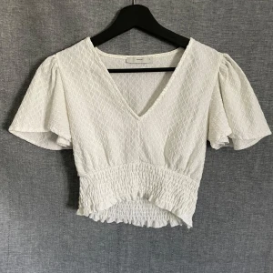 Vit croppad blus med volang - Säljer en söt vit croppad blus med v-ringning och korta, vida ärmar. Blusen har smockad midja och volangdetalj nedtill, vilket ger en luftig och somrig känsla.