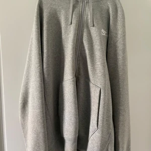 Grå hoodie med dragkedja från Nike - Säljer en klassisk grå hoodie från Nike med dragkedja och huva. Tröjan har fickor framtill och en liten vit Nike-logga på bröstet. Perfekt för en avslappnad stil och enkel att matcha med det mesta.