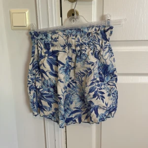 Blommiga shorts med blått mönster - Säljer ett par luftiga shorts med blått blommönster och resår i midjan. De är tillverkade i en blandning av viskos och bomull, vilket gör dem mjuka och sköna. Perfekta för varma dagar när du vill ha något bekvämt och snyggt.