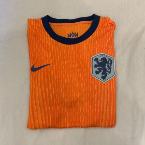 Fotbollströja storlek XL, Passar L - Säljer en officiell Holland fotbollströja från Nike med KNVB-emblem på bröstet. Tröjan är orange med tunna gula ränder storlek XL men passar L, har korta ärmar och svarta detaljer vid krage och ärmslut. Tillverkad i Dri-FIT material för bästa komfort.