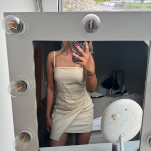 Beige omlottklänning med smala axelband - Säljer en beige linne kortklänning med smala axelband och omlottdetalj framtill. Klänningen har en rak, enkel passform. Helt oanvänd 