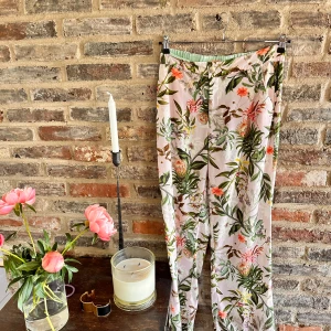Blommiga byxor med knäppning - Säljer ett par lätta byxor med blommigt mönster i grönt, rosa och orange på ljus botten. Byxorna har normal passform, knapp och resår i midjan samt fickor bak. Perfekta för en färgglad look! Finns matchande kavaj till.