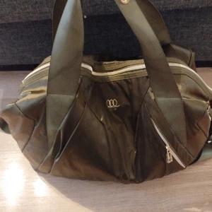 Olivgrön axelväska från Björn Borg - Säljer en olivgrön axelväska från Rituals med breda handtag och justerbar axelrem. Väskan har dragkedja upptill, guldfärgade detaljer och en diskret logga framtill. Perfekt för gymmet eller som yogabag då mattan är perfekt att knäppa fast ovanpå väskan.