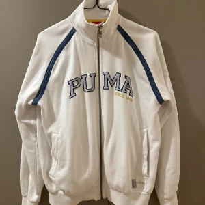 Vit vintage zip-jacka Puma - Vit kofta med dragkedja från Puma, har blå detaljer längs axlarna och broderad logga framtill. Hög krage och två fickor på sidorna. Vintage stil