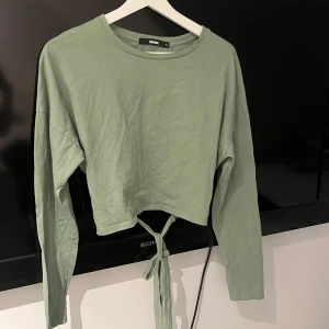 Grön croppad långärmad topp med knytning - Snygg ljusgrön croppad topp från BikBok med långa ärmar och knytband i midjan. Perfekt för en trendig och avslappnad stil. Mjuk bomullskänsla och enkel rund halsringning.