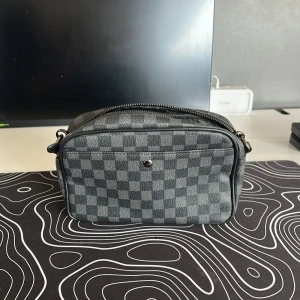 Svart rutig axelväska - Säljer en svart axelväska med rutigt mönster och dragkedja upptill. Väskan har en kompakt form och är tillverkad i syntetmaterial. Perfekt för att bära med sig det viktigaste.