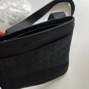 Svart axelväska från Emporio Armani - Snygg svart axelväska från Emporio Armani med diskret geometriskt mönster och logotyp framtill. Väskan har justerbar axelrem och dragkedja upptill. Perfekt för att bära med sig det viktigaste i stil. Ny pris 2300