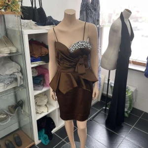 Brun kort klänning med strass och rosett - Snygg brun kort klänning med smala axelband och glittrande strassdetaljer över bysten. Klänningen har en markerad midja med stor rosett och peplumdetalj som ger en festlig look. Perfekt för dig som vill sticka ut. Finns i storlek 36-42, erbjuder även att skräddarsy efter dina egna preferenser!🥰