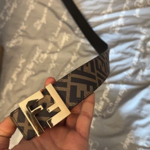 Fendi bälte  - Säljer ett snyggt bälte från Fendi med klassiskt FF-mönster i brunt och svart. Bältet har ett stort guldfärgat F-spänne och är gjort i skinn. Perfekt accessoar för att lyfta din outfit.