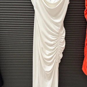 Vit långklänning med drapering och smala axelband - Säljer en elegant vit långklänning med smala axelband och snygg drapering längs sidan. Klänningen har en cowlneck-ringning och är tillverkad i ett mjukt, följsamt material som ger en lyxig känsla.