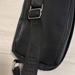 Svart axelväska från Burberry - Snygg svart axelväska från Burberry med justerbar axelrem och diskret logga på remmen. Väskan har dragkedja och detaljer i metall. Perfekt för att bära det viktigaste med stil.                   Säljer den för 1500kr men priset kan diskuteras! 