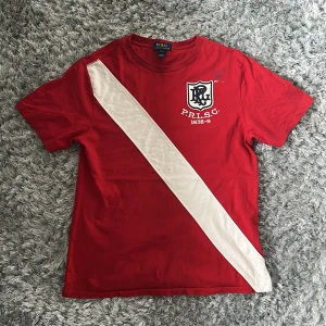 Röd t-shirt med vit diagonal rand från Polo Ralph Lauren - Säljer en röd t-shirt från Polo Ralph Lauren med en bred vit diagonal rand över framsidan och broderad logga på bröstet. Klassisk rund hals och korta ärmar. Perfekt för en sportig och stilren look.