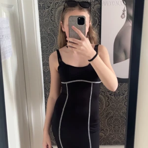 Svart tight klänning med kontrastsömmar - Svart tight klänning med vita kontrastsömmar och breda axelband. Klänningen har en kroppsnära passform och är ärmlös, perfekt för en trendig och stilren look.