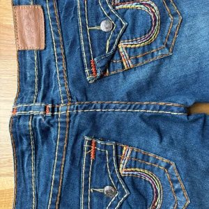 Blå jeansbyxor från True Religion med färgglada sömmar - Säljer ett par blå jeansbyxor från True Religion med jättesnygga fickor och detaljer. Säljer dem då de inte passar. De är storlek 27