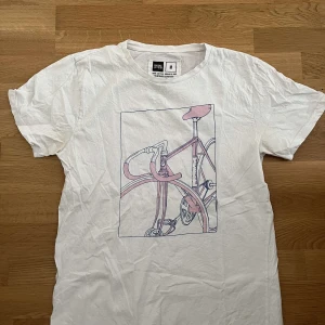 Vit t-shirt med cykelmotiv - Vit t-shirt från Junk de Luxe i bomull med tryck av en rosa och grå cykel på bröstet. Klassisk rund hals och korta ärmar. Perfekt för dig som gillar unika och kreativa prints.