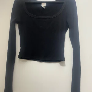 Svart ribbad långärmad topp H&M - En svart, ribbad långärmad topp från H&M med rundad, bred urringning. Modellen är figurnära och croppad, perfekt att styla med höga jeans eller kjol. Mjuk och stretchig i bomullsmix för en bekväm känsla hela dagen.