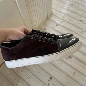 Svarta sneakers från Lanvin i mocka och skinn - Säljer ett par svarta sneakers från Lanvin med ovandel i mocka och tåparti i skinn. Skorna har snörning, vit platt sula och rund tå. Perfekta för dig som gillar stilrena och klassiska sneakers med en lyxig touch. Skorna är helt nya. 