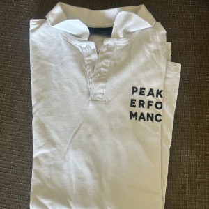 Vit Peak Performance pikétröja storlek XL - Snygg vit pikétröja från Peak Performance med svart logotyptryck på bröstet och en svart detalj på ärmen. Klassisk krage, kort ärm och knappar framtill. Perfekt för en clean och sportig stil. Tillverkad i mjuk bomull för skön känsla.