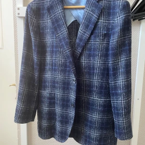 Oscar Jacobsson  kavaj - Snygg blå kavaj från Oscar Jacobsson i regular fit-modell med rutigt mönster. Tillverkad i en blandning av linne, ull och polyester, fodrad med viskos och polyester. Klassisk design med två fickor fram och en bröstficka. Perfekt för dig som gillar stilrena detaljer. Något uppsydda i ärmarna 