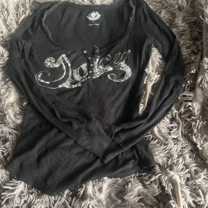 Juicy Couture svart topp med paljetter - Svart ribbad långärmad topp från Juicy Couture med djup rund halsringning och knappar framtill. Stor silverpaljett-text 'Juicy' på bröstet ger en extra glittrig vibe. Perfekt för dig som gillar statement-plagg och vill sticka ut.