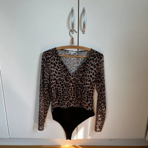 Leopardmönstrad meshbody från Amisu - Säljer en snygg långärmad body från Amisu med leopardmönster i beige och svart. Toppen har v-ringning och är gjord i tunn mesh. Storlek L . 