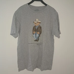 Grå Polo Bear t-shirt från Ralph Lauren - Snygg grå t-shirt från Polo Ralph Lauren med ikoniska Polo Bear-trycket i cowboy-outfit på bröstet. Klassisk passform med rund hals och korta ärmar. Perfekt för sommarens varma dagar. Små defekter på björnen
