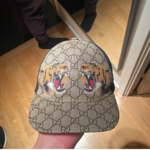 Gucci keps beige - Snygg keps från Gucci med klassiskt GG-mönster i beige och brunt framtill. Baksidan är i svart mesh för extra ventilation och justerbar rem med grönt och rött band. Märkesetikett på insidan och exklusiv känsla. Perfekt för dig som vill sticka ut med stil. Jättebra skick