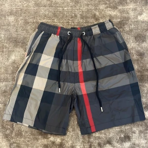 Burberry badshorts  - Snygga badshorts med den klassiska rutigt Burberry mönster i svart, grått, beige och rött. Shortsen har elastisk midja med snörning och en bakficka. Perfekta för sommaren och strandhäng. Priset är ej hugget i sten vid snabb affär!