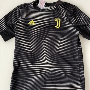 Juventus Adidas svart matchtröja S - Juventus fotbollströja från Adidas i svart med vita ränder och gula detaljer. Tröjan har korta ärmar, modern design och är tillverkad i lätt, återvunnet material (Parley/Climalite). Perfekt för dig som älskar fotboll och vill sticka ut på planen.