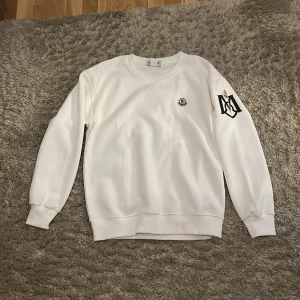 Vit Moncler sweatshirt med logga - Vit sweatshirt från Moncler med rund hals och långa ärmar. Tröjan har Moncler-logga på bröstet och en stor svart M-logga på ena ärmen. Ribbstickade muddar vid ärmslut och nederkant. Perfekt för en clean och stilren look helt ny priset kan diskuteras