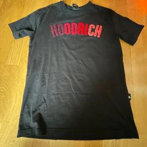Svart HOODRICH t-shirt med röd text - Svart t-shirt från Hoodrich i storlek XS med fet broderad HOODRICH-text i rött och vinrött på bröstet. Klassisk passform med korta ärmar och rund hals. Perfekt för dig som gillar streetwear och vill sticka ut med en snygg logga. Pris kan diskuteras