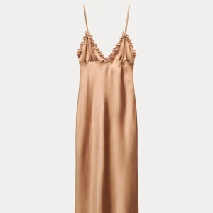  långklänning - Snygg långklänning i champagnefärgad satin med smala axelband och volangdetaljer vid bysten. Klänningen har en elegant, slät siluett och djup v-ringning. Perfekt för dig som gillar en stilren och trendig look. Köptes för 499