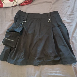 Svart kortkjol med avtagbar ficka - Svart kortkjol med coola detaljer som D-ringar och en stor avtagbar ficka med dragkedja framtill. Kjolen har bred linning och dragkedja bak. Perfekt för dig som gillar streetstyle och vill ha något unikt i garderoben.