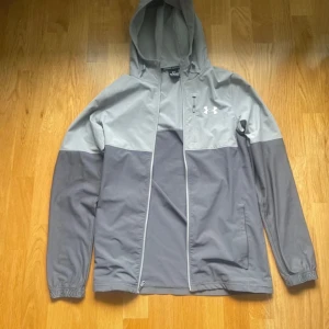 Grå vindjacka från Under Armour XS - Snygg tvåfärgad grå vindjacka från Under Armour i storlek XS. Jackan har huva, dragkedja framtill och elastiska muddar med logga på ärmsluten. Perfekt för blåsiga dagar och sportiga outfits.