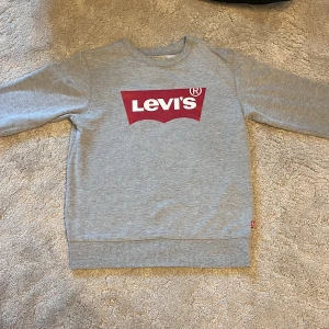 Grå sweatshirt från Levi's med logga - Säljer en grå sweatshirt från Levi's med den klassiska röda loggan framtill. Tröjan har rund hals, långa ärmar och ribbade muddar vid ärmslut och nederkant. Perfekt för en chill och stilren look.