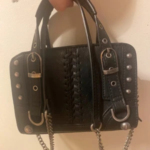 Svart Zara väska med nitar - Cool svart handväska i skinn med silverfärgade nitar, spännen och kedjerem. Väskan har dekorativ snörning framtill och detaljerade remmar med metallringar. Perfekt accessoar för en edgy look och rymmer det viktigaste du behöver ha med dig.
