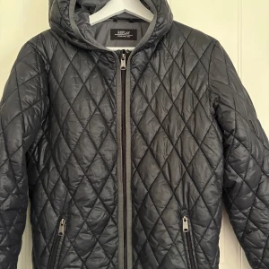Svart quiltad jacka från Replay - Quiltad svart jacka från Replay med huva och dragkedja framtill. Jackan har ett snyggt rutmönster, två fickor med dragkedja och är tillverkad i lätt syntetmaterial. Perfekt för kyliga dagar när du vill ha en stilren look.