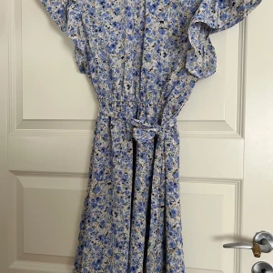 Blommig långklänning från SHEIN - Långklänning från SHEIN med blått och vitt blommönster, volangärmar och knytskärp i midjan. Lätt och flowy modell, perfekt för varma dagar. Snygg v-ringning och avslappnad passform som ger en romantisk vibe.