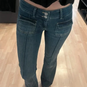 Blå bootcut jeans med fickdetaljer - Snygga blå jeans med bootcut passform och dubbla knappar i midjan. Jeansen har markerade sömmar längs benen och klassiska bakfickor. Perfekta för dig som gillar en trendig och avslappnad stil.