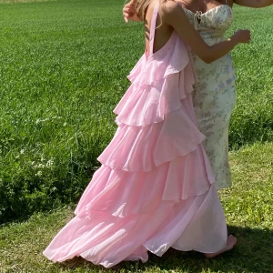 Ljusrosa volangklänning från Vero Moda - Drömmig långklänning i ljusrosa från Vero Moda med flera lager volanger och öppen rygg. Klänningen har breda axelband och en romantisk rosettdetalj framtill. Perfekt för sommarens speciella tillfällen. Lite trasig på kanten längst ner på klänningen men inget man tänker på då den har såpass mycket volanger.