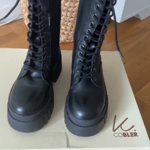 Svarta boots från K.Cobler med platåsula - Tuffa svarta boots från K.Cobler med grov platåsula och snörning hela vägen upp. Skorna är i skinn med textilfoder och har dragkedja på insidan för enkel påtagning. Perfekta för dig som vill ha en edgy look till höst och vinter. Har inte använt den, nypris 1500kr säljer för 900kr
