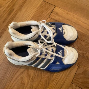 Adidas inomhus träningsskor blå och vita strl 38 - Snygga Adidas inomhus träningsskor i vitt och blått med klassiska tre ränder på sidan. Skorna har snörning och kardborreband framtill, samt slitstark gummisula i orange. Perfekta för dig som gillar inomhus sport som futsal eller innebandy! Använda en del, pris kan diskuteras!