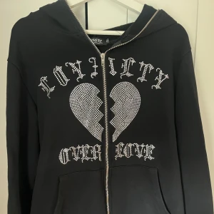 Svart hoodie med tryck i rhinestone - Svart hoodie från Trotherz med dragkedja, huva och stora rhinestone detaljer framtill med texten 'Loyalty Over Love'
